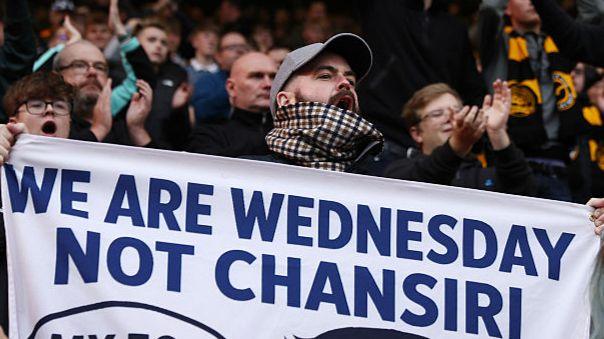 A football fan in a grey baseball cap and blue and brown check scarve shouts among other fans as he holds up a banner which reads "We are Wednesday, not Chansiri"