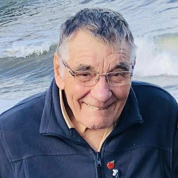 An older man stands in front of the sea, waves in the background. He wears glasses, a navy zip jacket with a red poppy pin, and has short gray hair. He is smiling at the camera.