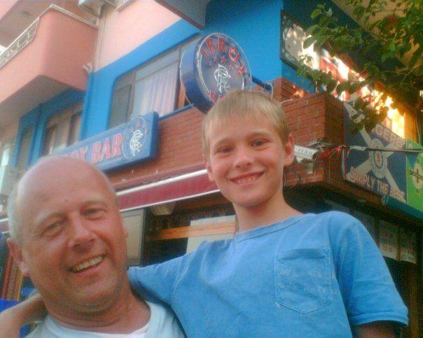 Brian Innes has his young son in his arms while on holiday. They are tanned and smiling and both have light blue tops on.