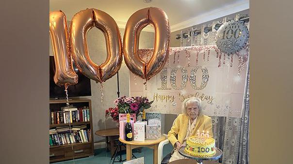 Elderly woman in a yellow blouse and jacket wears cream slacks and cream flat shoes. She is seated and in front of her is a large yellow and cream-iced birthday cake with 100 in piped icing on it. On another table are cards, champagne and balloons and a banner with 100 on them.