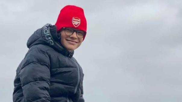 Harry Basham is sitting on a rock on a beach. He is wearing a red hat and a black hooded jacket. He is wearing black tracksuit bottoms and black and red trainers. The picture was taken on a cloudy day.