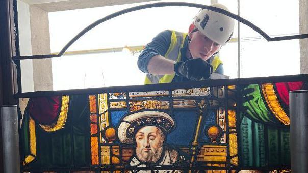 A large and colourful stained glass panel which shows Henry VIII handing a charter to Sir Thomas Vicary. The panel is full of rich yellows, blues and reds. Behind the panel is a man wearing a hard hat and high vis jacket, appearing to fix the top of the panel. It is unclear what tool he is using.