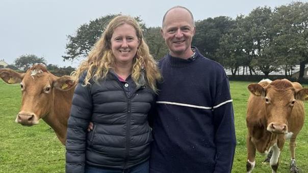 Lady with blonde curly hair in a navy puffer jacket stood to the left of a bald man in a navy knitted jumper smiling at the camera. There is a brown cow next to them looking at the camera.