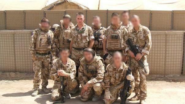 Two rows of soldiers, some armed with rifles, posing for a photo wearing desert camouflage uniform with a sand-coloured protective wall behind them. Nine of the ten faces have been blurred and only Chris Draper is identified