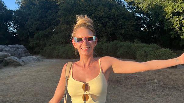 Rhiannon Quinche stands outdoors at Tunbridge Wells Common in Kent wearing eclipse glasses. Trees, dry grass and large rocks can be seen behind her in the evening sunlight.