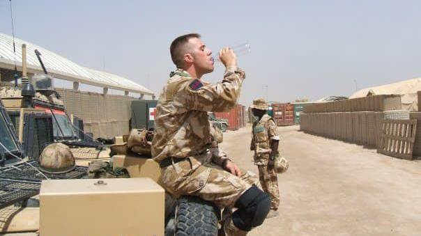 Solider Chris Draper wearing desert camouflage uniform, sat on the tyre on the front of a military 4x4 vehicle while drinking from a water bottle in a Middle Eastern military camp with shipping containers in the background. 