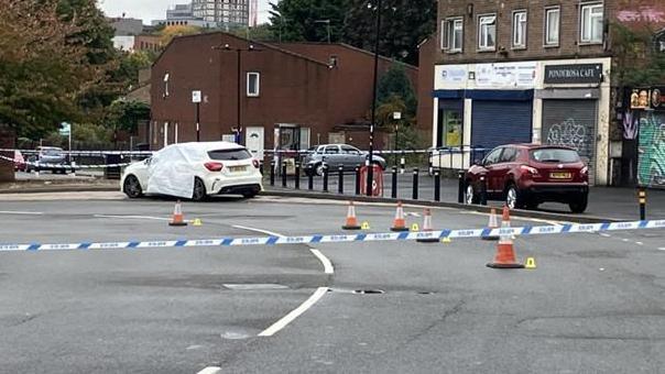 A cordoned-off street. A white car is covered with a protective sheet, suggesting it may be central to the incident. Nearby, a red car is parked, and the area is marked off with police tape and traffic cones to restrict access. The surroundings include buildings with signage and graffiti art, and in the distance, a crane and tall structures indicate a city environment.