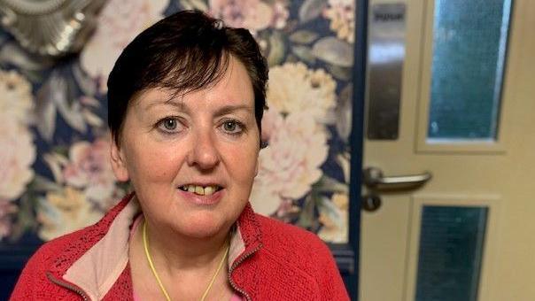 Fiona Mulhern is looking at the camera. She had a red top on and has dark hair. The background is flowery wallpaper and a door.