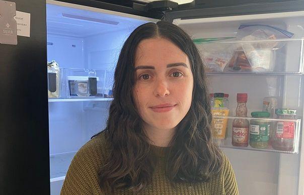 Jessica Ireland has black wavy hair and is standing in front of a fridge which has an open door and condiments in the background.