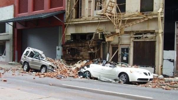 A white car and a silver car completely destroyed on one side by fallen bricks and debris. The front of the buildings behind them are damaged with railings falling off. 