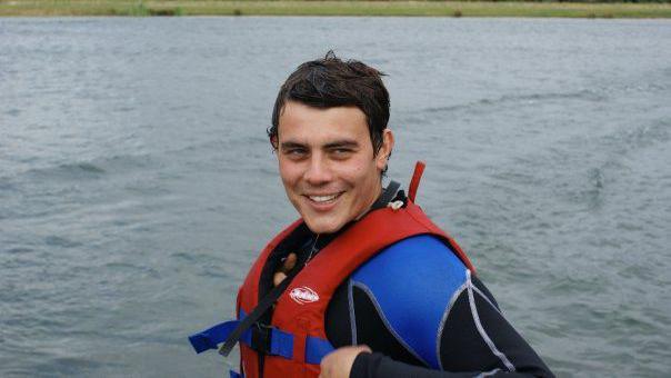 Robert Chapman has short brown hair and is smiling. He is wearing a blue and black wetsuit and a red life jacket. He is surrounded by water.