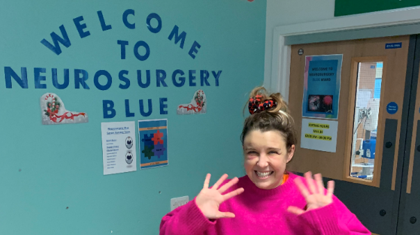 Ms Wrathall smiles at the camera with her hands near her face in a wave. She wears a bright pink jumper. Behind her on a light blue wall, navy letters read: Welcome to Neurosurgery Blue.