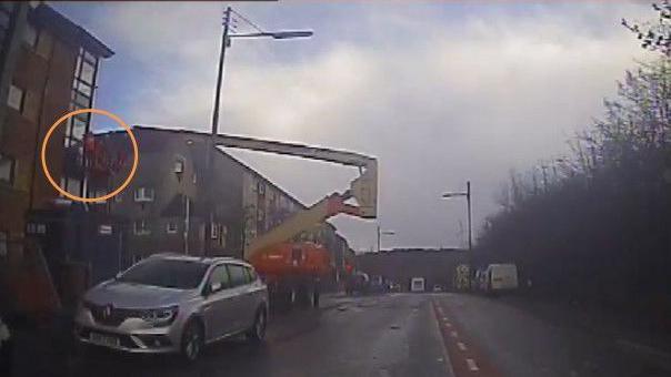 CCTV dated 04/02/22 issued by the Crown Office & Procurator Fiscal Service (COPFS) showing a roofer (circled) before he was thrown from a cherry picker on a street with flats along the lefthand side in Castlemilk, Glasgow. There is a silver Renault car in the foreground.