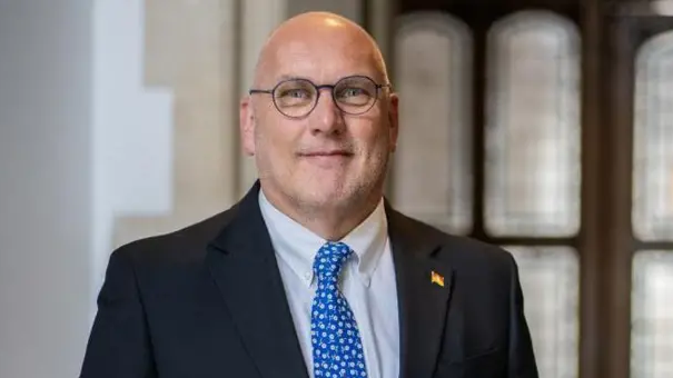 Nick Adams-King, who is bald, wearing a spotted blue and white tie and a black jacket.