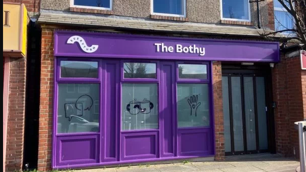 The Bothy is a purple fronted building with a door to one side and three windows, one with various drawings like a hand in the air, and two cups of tea 