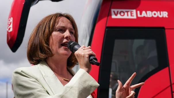 Lola McEvoy speaking into a microphone at an election campaign event in 2024.  Behind her is a red bus with "Vote Labour" branding on the side.