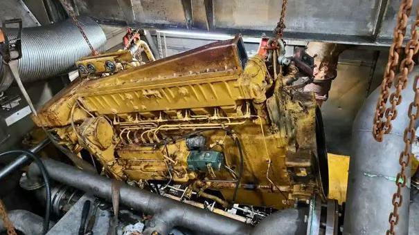 A large yellow-coloured metal engine, with many wires and sections of different shapes is hoisted out of a tight space in a grey metal area of the vessel's engine.