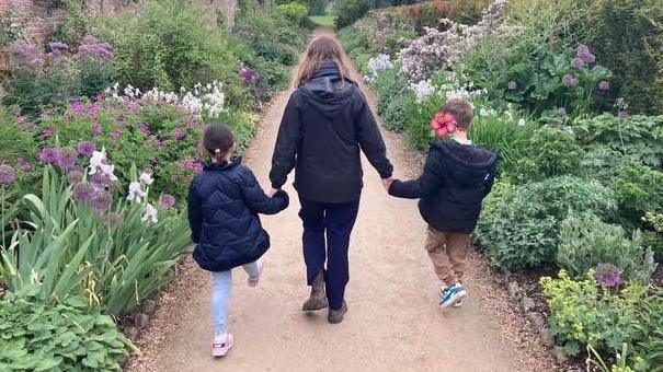 A woman in a blue coat holds hands with a boy and girl as they walk through a formal garden. All have their backs to the camera.