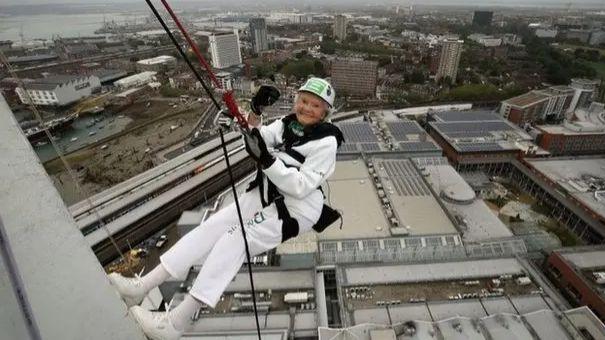 A 101-year-old woman named Doris Long abseiling down the side of the Spinnaker Tower. She is equipped with safety gear, including a helmet and harness, and is smiling toward the camera. In the background, Portsmouth can be seen. There is a wide urban landscape featuring buildings, roads, and patches of greenery, offering a panoramic view from a high vantage point.