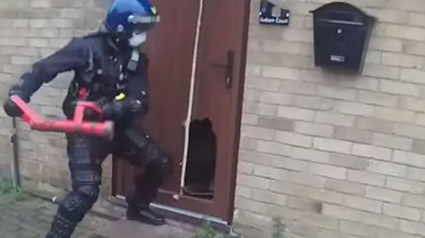 A police officer with a red smashing their way into a brown door at a brick house