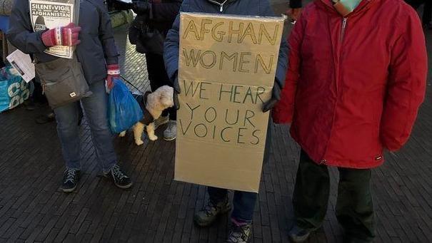 Seven campaigners outside Next department store in Norwich. Two women, both in blue coats and hats, are holding signs and placards. One reads 'Afghan women we hear your voices'. A lady in a red coat and short white hair is stood alongside the women