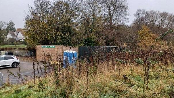 A large wooden shed and a shipping container can be seen at the edge of a car park. Some cars are parked nearby and there are shrubs and trees around the area, with some houses also visible.