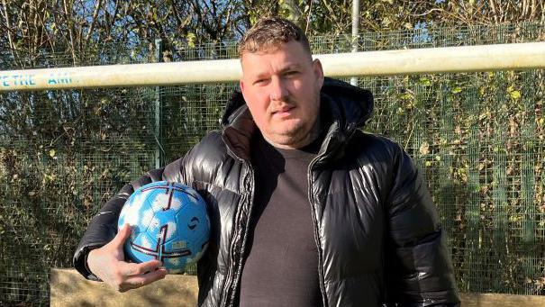 Charlie Barker is standing in front of a football goal. He is wearing a black t-shirt, black puffer jacket and is holding a blue football under one arm.