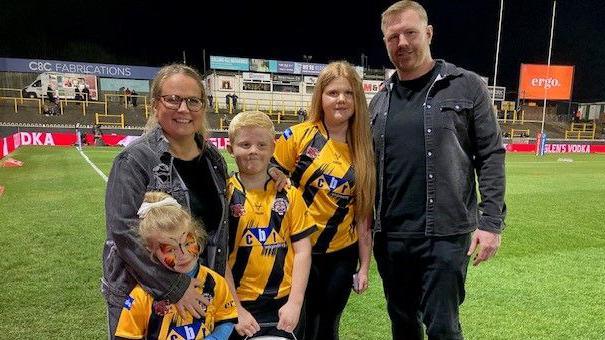 A mum and dad stand with their three young children on a floodlit sports pitch. The children - two girls and one boy - are all wearing and yellow and black sports shirts. The parents are wearing dark denim jackets and black t-shirts.