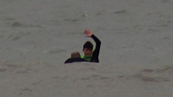 Oscar is in a grey sea signalling with his arm over his head and the head and shoulders of a man is visible in the water next to him.