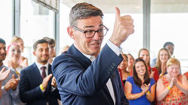 Andy Burnham gives a thumbs up as he stands in front of a clapping crowd of supporters.