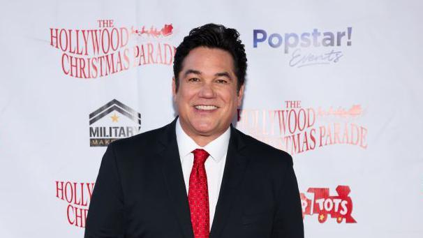 Dean Cain pictured in 2024 at a Hollywood Christmas Parade red carpet wearing a black suit, white shirt and red tie.