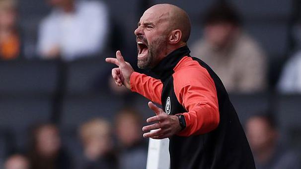 MK Dons boss Paul Warne in animated pose, shouting from the sidelines with his arms outstretched