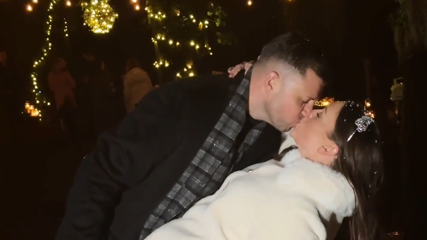 Michael and Sinead Lysaght kiss after marrying in a forest in December. She is wearing a white coat and silver hairband and he is wearing a dark coat with grey checked scarf. 