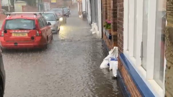 Cars in heavy flash flooding alongside a row of terraced houses with sandbags in the windows