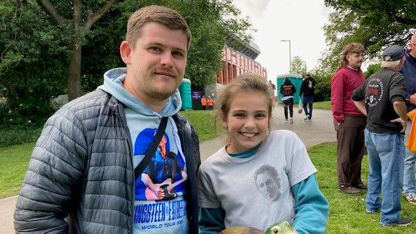 Ollie has short hair and a short beard and is wearing a blue Bruce Springsteen hoodie and quilted jacket. Mollie is wearing a light blue Bruce Springsteen t-shirt over a darker blue long sleeved top.