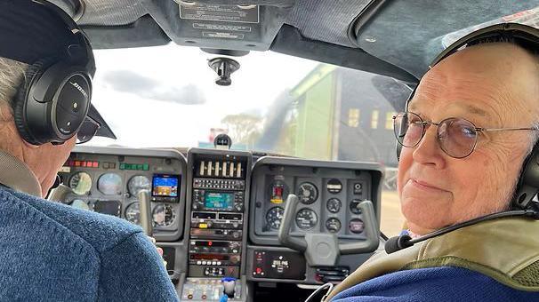 Two men sitting in the cockpit of a Cessna light aircraft. On the right is Jack Waterfall, 75, with a bald head and wearing glasses and a blue jumper with a khaki sleeveless jacket on top. Both men are wearing headphones. The pilot is the man on the left, wearing a blue wool jumper. He is looking ahead at the controls and his face is not visible