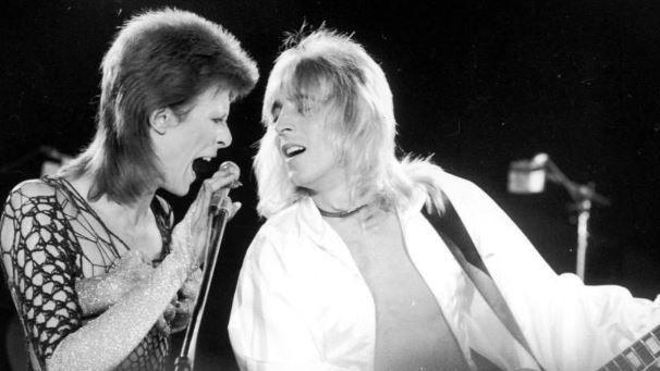 A black and white photo of Bowie with his mouth open singing into a microphone. He is wearing a netted top and looking at Ronson who is wearing an open white shirt and looking back at him, while playing guitar