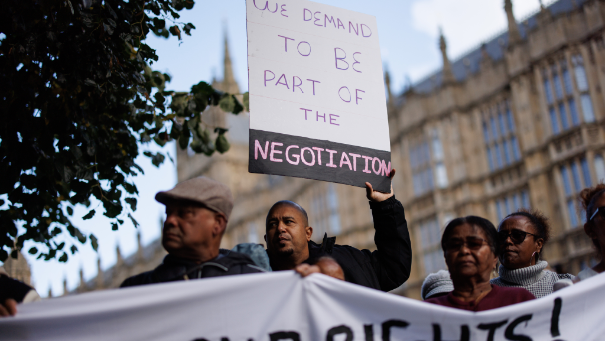 A group of people standing outside the Houses of Parliament. One holds a sign that reads: "We demand to be part of the negotiation."