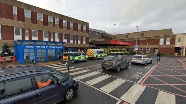 Car drop-off point in front of a low, brown building with a blue sign reading: Diana, Princess of Wales Hospital