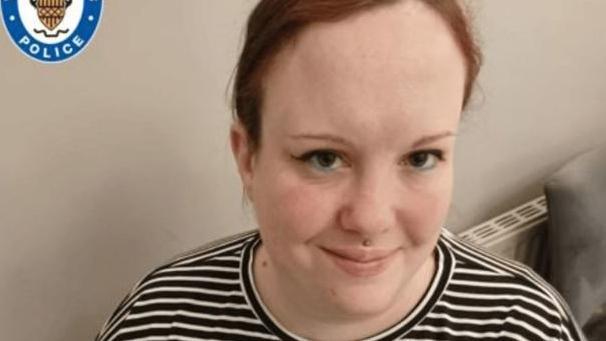 A young woman with red hair, tied back, and brown eyes smiles into the camera. She has eyeliner which flicks at the side and is wearing a stripey t-shirt