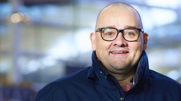 Jeremy Smile smiles at the camera. He is bald and wears glasses with a navy coloured coat.