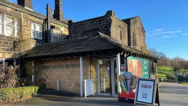 The front entrance of Abbey House Museum, a brown, Victorian-style brick building set in green gardens, with signs and ice cream advertisements outside.