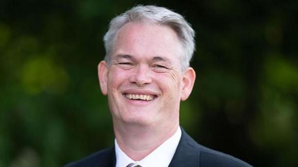 A head and shoulders photograph of Richard Tunnicliffe, wearing a black suit and a white shirt. He has grey hair and is smiling into the camera. Greenery is in the background and is out of focus.
