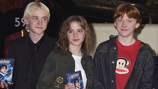 Tom Felton, Emma Watson and Rupert Grint, pictured as children, standing beside each other holding DVDs of Harry Potter and the Philosopher's Stone.