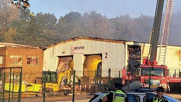 A white corrugated metal warehouse following the fire. The roof is burnt and still smouldering. 