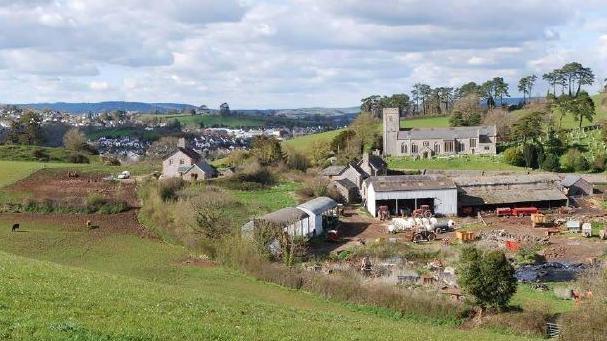 A view of part of the site. It is on green rolling countryside. An old church is in the middle of the picture. A farm is in the foreground with machinery and barns. Housing and trees sprawl off into the distance. 