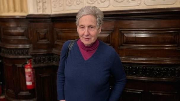 Jenny, with short grey hair, is looking straight at the camera in a wood-panelled room 