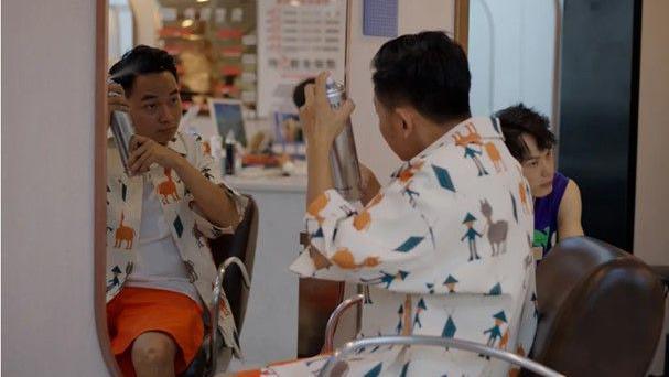 Hao sitting in front of a mirror spraying hairspray into his hair at the hairdressers