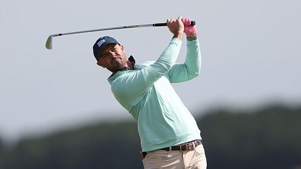 Wesley Bryan playing in The Open Final Qualifying wearing a navy blue cap and a turquoise long-sleeve top. Bryan is in the middle of his back swing after a hitting a shot with an iron.