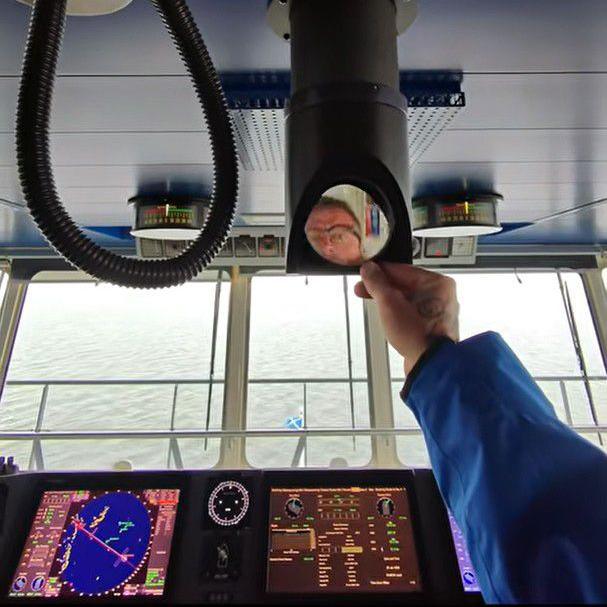 A man wearing glasses looks into a small mirror on a piece of equipment in the bridge of a large ferry